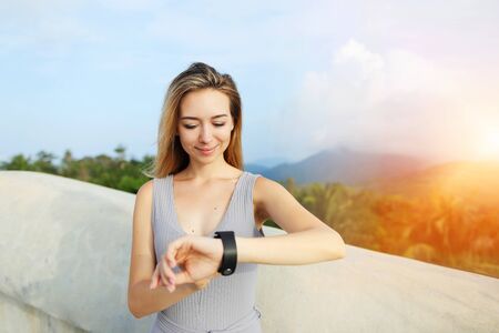 Sunshine portrait of young blonde woman using smartwatch, mountains in background, Thailand. Concept of tropical resort, modern technology and summer vacations.の写真素材