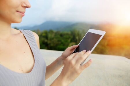 Bright photo with sunshine of blonde woman using tablet, mountains in background, Bali. Concept of tropical resort, modern technology and summer vacations.の写真素材