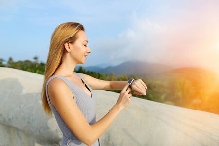 Sunshine portrait of young caucasian girl using smartwatch, mountains in background, Thailand. Concept of tropical resort, modern technology and summer vacations.の写真素材