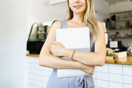 Blonde housewife standing with closed laptop in kitchen and smiling. Concept of happy wife and modern technology.の写真素材