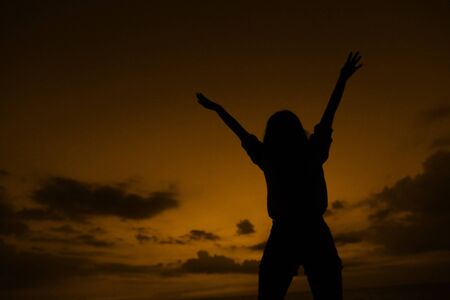 Black female silhouette with raised hands on brown sunset background with clouds in Thailand. Concept of cheap last minute tours to tropical resort, resting and freedom.の写真素材