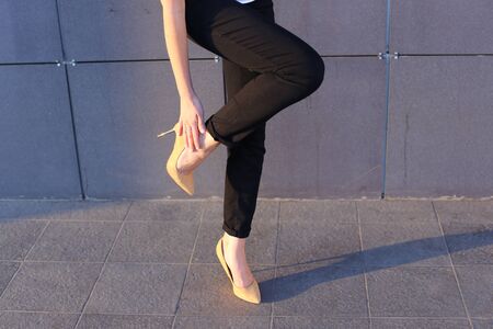 Girl holds leg rotated in profile at camera against background of gray wall of modern business center. Girl dressed in black classic trousers and beige high heels. Concept of advertising, advertising clothes, shooting lookbook, tired feet.の写真素材