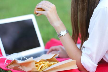 female in the park works behind laptop. beautiful girl with glasses eating fast food in nature and smiling.の写真素材