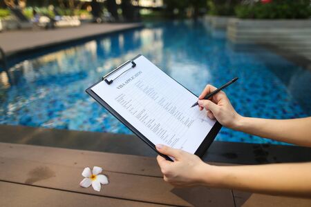 female hand holds out pen, fills out application form in United States. Sunny portrait on pool background And sunlight. concept of traveling to new countries. Front used with Open Font Licenseの写真素材