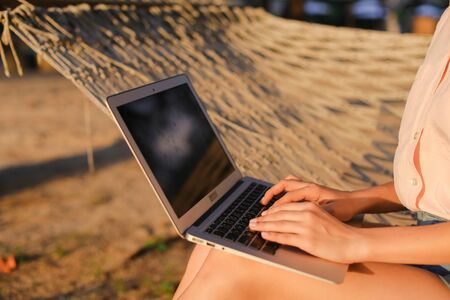 Close up female person using laptop and sitting on wicker hammock. Concept of modern technology and resting on beach.の写真素材