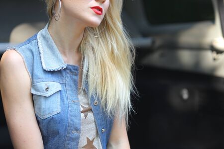Shooting of parts of girl's image, young girl sitting in blue car SUV jeep. On lips of girl red lipstick, also photo shows wear, T-shirt with stars, beige shorts and denim vest with pockets of accessories on girl gray round earrings and lies next to small blue bag.Concept of shot of details of clothing, advertising, travel, automobile.の写真素材
