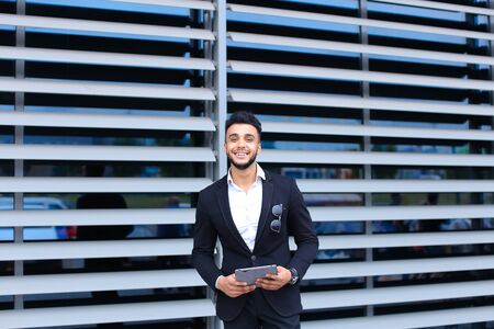 Young handsome businessman Arab Muslims is in the business center in black elegant dress on building background. Businessman man male looking waiting concept and smiling.の写真素材