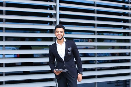 Young handsome businessman Arab Muslims is in the business center in black elegant dress on building background. Businessman man male looking waiting concept and smiling.の写真素材