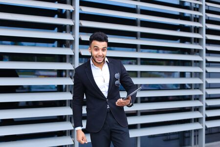 Young handsome businessman Arab Muslims is in the business center in black elegant dress on building background. Businessman man male looking waiting concept and smiling.の写真素材