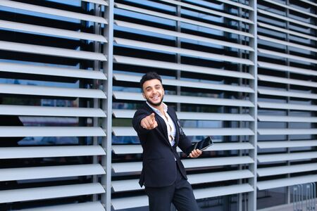 Young handsome businessman Arab Muslims is in the business center in black elegant dress on building background. Businessman man male looking waiting concept and smiling.の写真素材