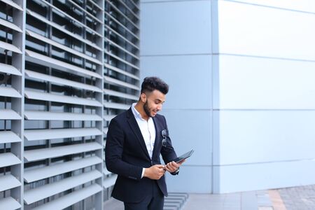 Young handsome businessman Arab Muslims is in the business center in black elegant dress on building background. Businessman man male looking waiting concept and smiling.の写真素材