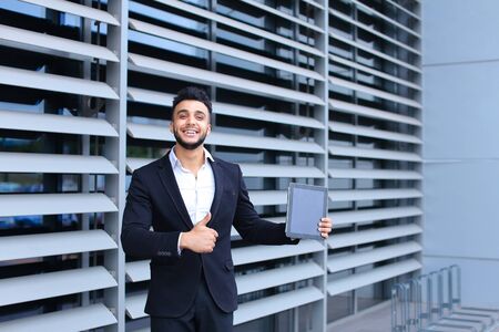 Young handsome businessman Arab Muslims is in the business center in black elegant dress on building background. Businessman man male looking waiting concept and smiling.の写真素材