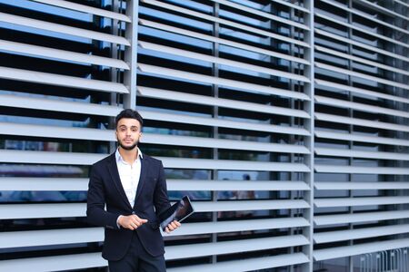 Young handsome businessman Arab Muslims is in the business center in black elegant dress on building background. Businessman man male looking waiting concept and smiling.の写真素材