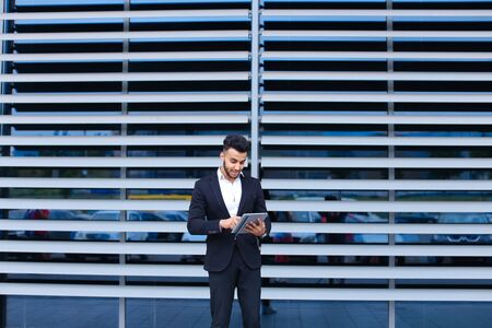 Young handsome businessman Arab Muslims is in the business center in black elegant dress on building background. Businessman man male looking waiting concept and smiling.の写真素材