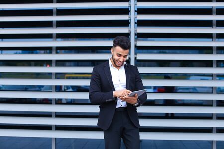 Entrepreneur uses tablet for online order shopping with debit credit card looking smiling at the camera. Young handsome businessman arab muslims in business center wearing dressed in black elegant suit on building background.の写真素材