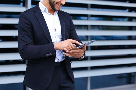 Entrepreneur uses tablet for online order shopping with debit credit card looking smiling at the camera. Young handsome businessman arab muslims in business center wearing dressed in black elegant suit on building background.の写真素材