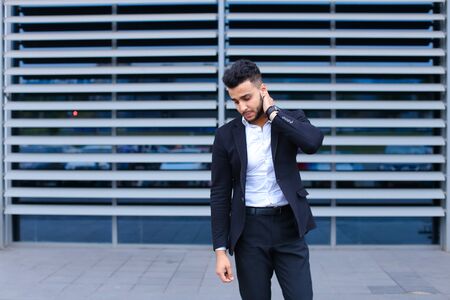 Entrepreneur shows neck pain tired looking at the camera. Young handsome businessman arab muslims in business center wearing dressed in black elegant suit on building background.の写真素材