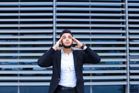 Entrepreneur shows neck pain tired looking at the camera. Young handsome businessman arab muslims in business center wearing dressed in black elegant suit on building background.の写真素材