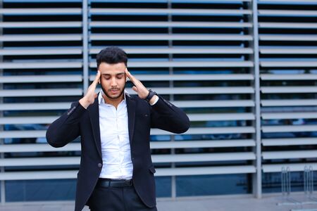 Entrepreneur shows neck pain tired looking at the camera. Young handsome businessman arab muslims in business center wearing dressed in black elegant suit on building background.の写真素材
