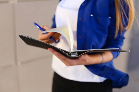 Girl holds in hands diary, black notebook with sheets and blue pen and leafs through pages. Young woman with light brown hair dressed in white T-shirt and bright blue shirt with pocket on arm bracelet.Concept of organization and planning of day, notebook, homework, busy dealer.の写真素材