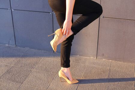 Girl holds leg rotated in profile at camera against background of gray wall of modern business center. Girl dressed in black classic trousers and beige high heels. Concept of advertising, advertising clothes, shooting lookbook, tired feet.の写真素材