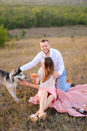 Young caucasian woman and man sitting on pink plaid near husky and resting outside. Concept of picnic on nature and dogs.の写真素材