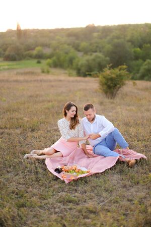 Happy caucasian woman and man sitting on pink plaid in field and resting. Concept of picnic on nature and relationship.の写真素材