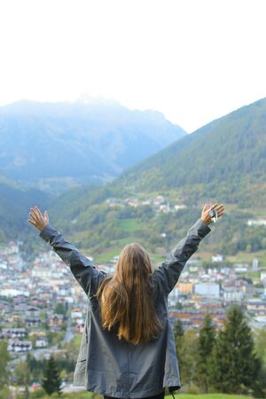 Girl with raised hands standing in mountains, buildings in background. Concept of mountain village, town and freedom.の写真素材