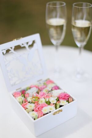 Two golden rings lying in flower white box near wineglasses with champagne. Concept of wedding decor elementsand celebrating marriage.の写真素材