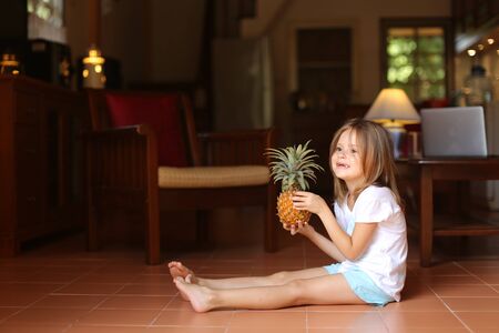 Little cute female kid sitting on floor in living room and playing with pineapple, laptop in background. Concept of health life, fruit and childhood.の写真素材