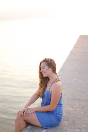 Young woman sitting on pier and wearing blue sundress. Concept of morning meditation in open air and relaxing near sea.の写真素材
