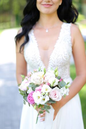 Brunette bride keeping bouquet of roses and wearing white dress. Concept of celebrating wedding and open air photosession.の写真素材