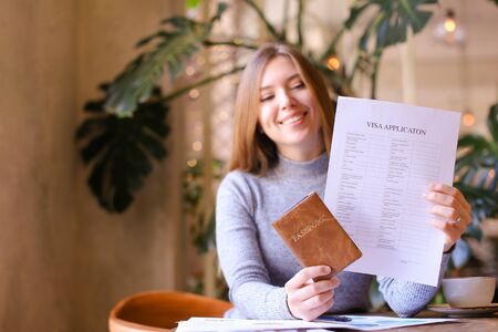 Young caucasian woman holding passport and visa application, sitting at cabinet, monstera in background. Concept of adult citizen and execution of documents.の写真素材
