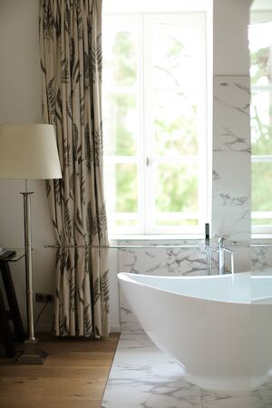 Modern white bathroom with marble wall and ceramic bath near window and floor lamp. Concept of home design and fashionable interior.の写真素材