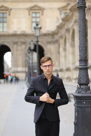 Young handsome man walking in Paris and leaning on lantern, weaaring black suit. Concept of walking in city and male fashion.の写真素材