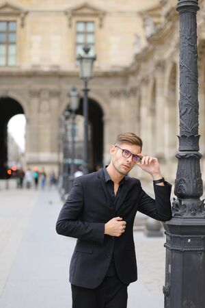 Young european man walking in Paris and leaning on lantern, weaaring black suit. Concept of walking in city and male fashion.の写真素材