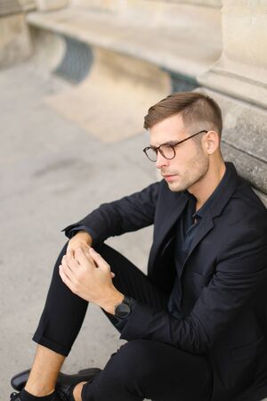 Young fashionable man sitting on sidewalk ground and wearing black suit, leaning on concrete banister. Concept of walking in cty and male stylish model.の写真素材