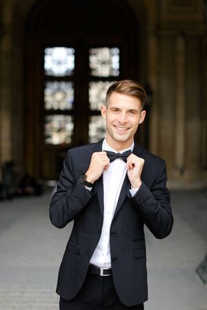 Young european man standing and posing, wearing black suit and bow tie, window in background. Concept of fashionable businessman and male model.の写真素材