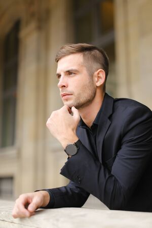Closeup portrait of young businessman on balcony leaning on banister. Concept of male model and fashion. Young man wearing black suit.の写真素材