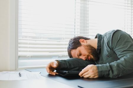 Exhausted bearded businessman sleeeping on workplacer, tablet on desk. Concept of financial crisis because of coronavirus and tired boss. Man using tablet and having brea for resting.の写真素材