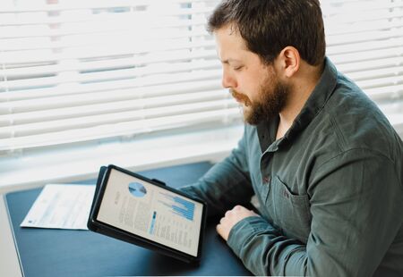 European male engineer working with graphs and diagrams on tablet at office. Concept of professional occupation and smart employee. Adult man surfing internet by modern gadget.の写真素材