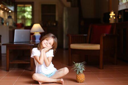 Little female kid sitting on floor in living room and keeping pineapple, laptop in background. Concept of health life, fruit and childhood.の写真素材