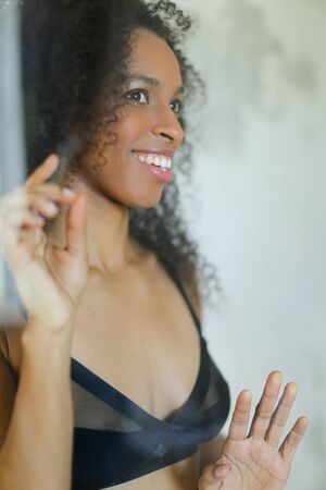 Portrait of young afro american smiling nice young woman wearing bra and touching glass in bathroom. Concept of underwear photo session.の写真素材