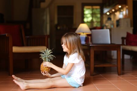 Little female blonde kid sitting on floor in living room and keeping pineapple, laptop in background. Concept of health life, fruit and childhood.の写真素材