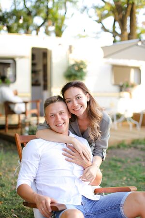 Young caucasian man and woman sitting in wooden chair near house on wheels. Concept of happy couple and summer vacations.の写真素材
