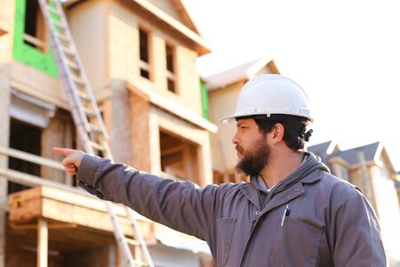 Adult foreman and engineer giving indication on construction site and wearinhg hardhat, brick building in backgound. Concept of builder profession and working abroad in Canada.の写真素材