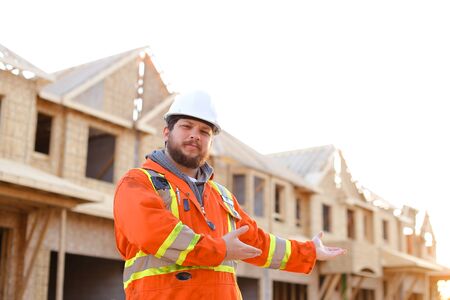 Male foreman wearing orange workwear and showing construction site, house in background. Concept of building new apartments in Canada and real estate.の写真素材