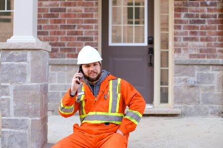 Foreman sitting in front of house and talkign by smartphone, wearing orange workwear. Concept of modern technology and builder profession.の写真素材