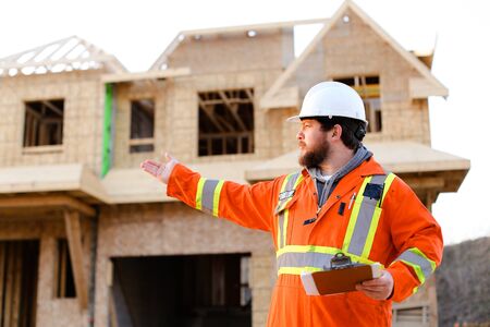 Foreman in orange workwear holding notebook on construction site, house in background. Concept of real estate in Canada.の写真素材