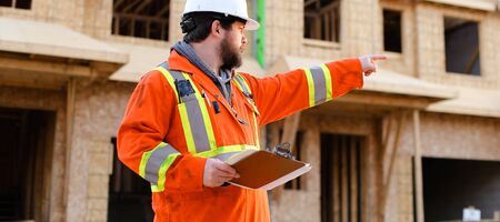 Banner of builder in orange workwear holding notebook on construction site, house in background. Concept of real estate in Canada.の写真素材